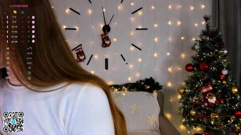 Media: Video of a woman with long hair, wearing a white top, facing a Christmas tree with ornaments and a clock background.