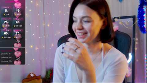 Media: Video of a happy, fair-skinned woman with shoulder-length brown hair, wearing a white blouse, seated in a chair with a blue cushion. Background features pink and purple fairy lights, a blue tinsel garland, and a Christmas tree.