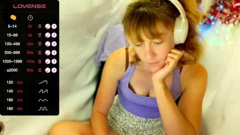 Media: Video of a young woman with shoulder-length brown hair, wearing a purple tank top and gray shorts, resting her head on her hand while listening to headphones in a cozy, warmly lit room.