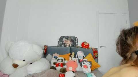 Video of a white room with plush toys and a framed picture on a bed; a person with wavy brown hair is partially visible.