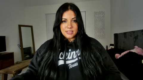 Media: Video of a middle-aged woman with long black hair, wearing a black hoodie, sitting in a cluttered bedroom with a bed, a mirror, and a TV.