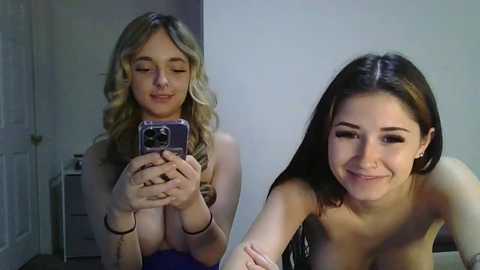 Media: Video of two young women, one taking a selfie with a phone, the other smiling, both topless, in a dimly lit room with white walls and a door.