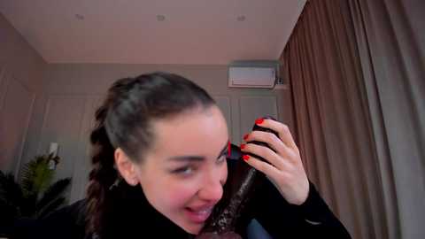 Media: Video of a young woman with fair skin and dark hair, laughing while holding a red phone to her ear. She wears a black coat and stands in a beige room with white doors, beige curtains, and a green plant in the background.