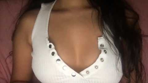 Media: Video of a woman with long, dark hair, wearing a white, ribbed tank top with button details, lying on a pink bed.