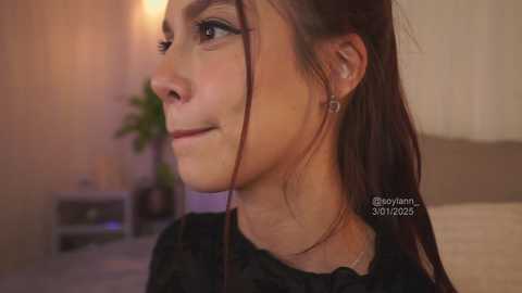 Media: Video of a young woman with light skin and brown hair, wearing black earrings and a black top, gazing thoughtfully. Background features a beige wall, a green plant, and a bed with white bedding.