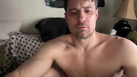 Media: A video of a shirtless man with a beard, light skin, and short brown hair, looking serious, sitting on a bed with a patterned duvet, black pillow, and a beige lamp beside him.