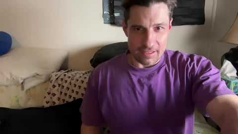 Media: Video of a man with short dark hair and a purple T-shirt, smiling slightly, in a cluttered living room with beige walls, a couch, and a lamp.