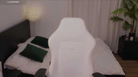 Media: Video of a modern bedroom featuring a white, ergonomic gaming chair, dark green pillows, a white fur rug, a tall white lamp, and a potted plant in the corner.