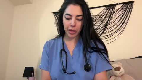 Media: A video of a young Latina woman with long black hair, wearing blue scrubs and a stethoscope, standing in a modern bedroom with a black macram\u00e9 wall hanging and a beige pillow on a bed.