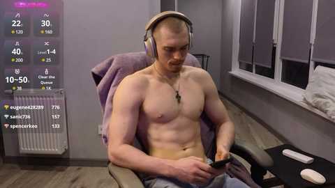 Media: Video of a muscular, shirtless, light-skinned man with a short beard, wearing headphones, sitting on a purple chair, looking at a smartphone. Background shows a modern room with gray walls, radiator, window blinds, and gaming equipment.