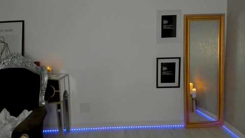 Media: Video of a minimalist bedroom with white walls, a bed with a black frame and silver bedhead, two framed black-and-white pictures, a tall golden-framed mirror, and blue LED lights under the wooden floor, creating a serene ambiance.