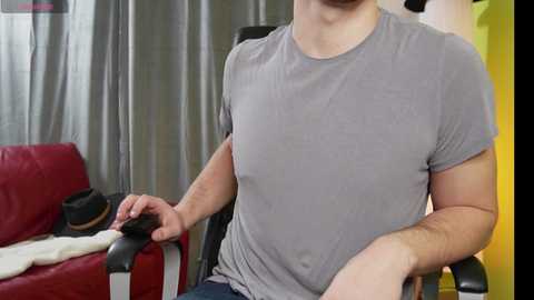 Media: Video of a light-skinned man with a medium build, wearing a gray t-shirt, sitting on a wheelchair in a living room. Background features a red couch, a yellow wall, and gray curtains.