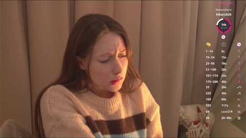 Video of a young Caucasian woman with long brown hair, wearing a beige and brown striped sweater, sitting indoors with a distressed expression, background includes a calendar overlay.