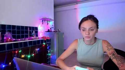 Media: Video of a woman with tattoos, light skin, and dark hair, wearing a light gray sleeveless top, seated in a kitchen with colorful Christmas lights, blue tiled backsplash, and a white wall.