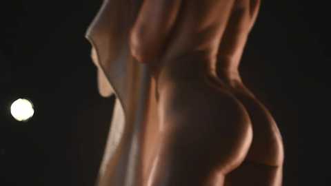 Media: A close-up video of a nude woman's backside, lit by a dim, warm light, revealing her smooth, tan skin. Her buttocks are prominent, and the background is dark, creating a stark contrast.