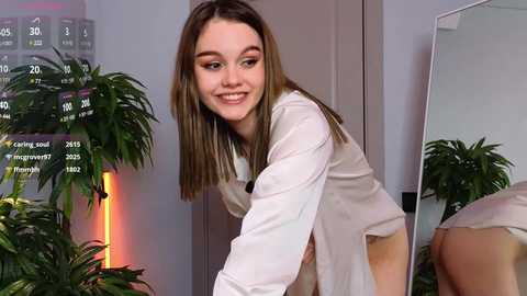 Media: Video of a young, smiling Caucasian woman with straight brown hair, wearing a white lab coat, bending over a table, surrounded by green plants and a calendar.