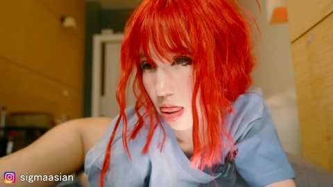 Video of a fair-skinned woman with bright red hair, wearing a light blue shirt, looking shocked or surprised indoors.