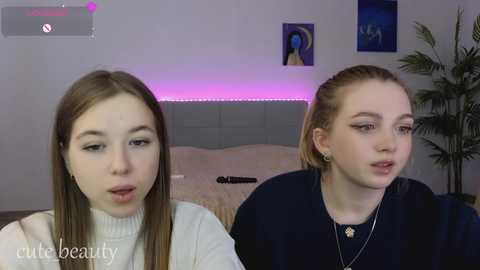 Video of two young women with light skin, straight brown hair, and fair complexion, wearing white and black tops, in a modern, dimly-lit bedroom with purple LED strip lighting and a potted plant.