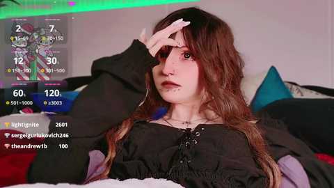 Media: Video of a young woman with long brown hair, wearing a black lace-up top, sitting on a bed with colorful pillows. She covers her eye with her hand. Live stream overlay shows chat messages and stats.