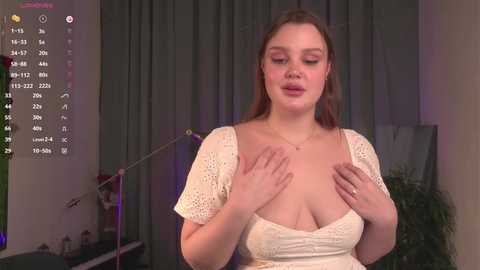 Media: Video of a fair-skinned, light-haired woman with large breasts, wearing a lacy white top, touching her chest, in a dimly lit room with a green curtain and potted plants.