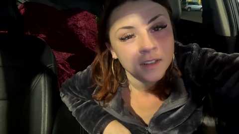 Media: Video of a light-skinned woman with brown hair, wearing a dark zip-up jacket, sitting in a car with red velvet upholstery. She has a neutral expression.