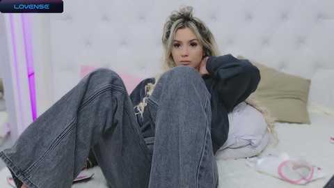 Media: Video of a blonde woman with long hair, wearing a black hoodie and denim overalls, lying on a bed in a white room with pink and purple lighting.