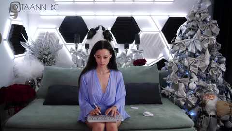 Media: Video of a woman in a blue robe, seated on a green couch, writing in a notebook, surrounded by festive decorations, including a Christmas tree, in a futuristic room.