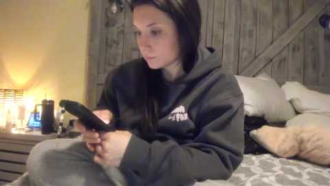 Media: Video of a young woman with long dark hair, wearing a dark hoodie, sitting on a bed, holding a smartphone, surrounded by pillows and wooden wall.