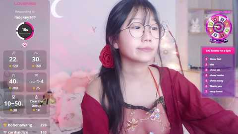 Media: Video of an East Asian woman with long black hair, glasses, and a red rose in her hair, wearing a red top, sitting indoors, with a video game interface overlay.