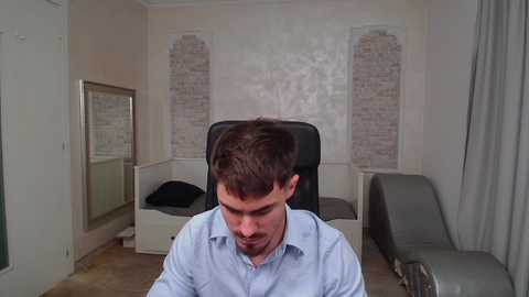 Media: A video of a man with short dark hair and a mustache, wearing a light blue button-down shirt, sitting in a modern, minimalist room with beige walls, a brick-patterned wall, a gray chaise lounge, and a black chair.