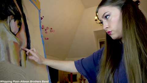 Media: Video of a young woman with long, straight brown hair, painting a detailed portrait of a woman's face on a canvas. She wears a dark blue shirt, focused intently on her work in a well-lit room.