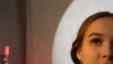 Media: Close-up video of a young woman with fair skin, brown hair, and green eyes, partially visible, in a modern bathroom with a round mirror.