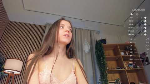 Media: Video of a young Caucasian woman with long brown hair, wearing a pink lace bra, standing in a dimly lit room with wooden shelves and vertical blinds.