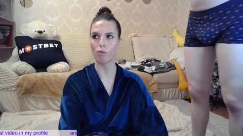 Media: Video of a woman with light skin and brown hair in a bun, wearing a blue robe, in a cozy living room with a plush bear, patterned wallpaper, and a man in boxer shorts.