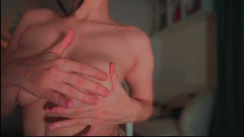 Media: Video of a topless woman with fair skin, medium-sized breasts, and a slender build. She has her hands covering her chest. The background is blurred, showing a bedroom setting.