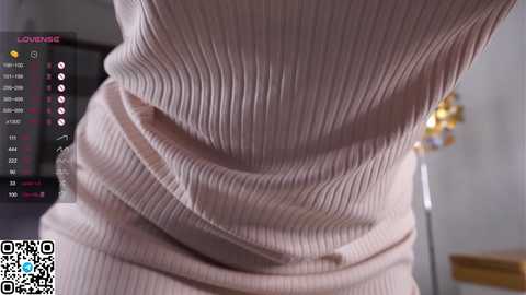 Media: A close-up video of a woman's waist and hips in a ribbed, light pink knit dress. The background includes a blurred living room with a TV screen displaying a \"Lover\" app.