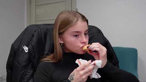 Media: A video of a young Caucasian woman with long, straight light brown hair, wearing a black turtleneck, eating a sandwich on a black Nike chair.