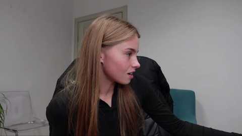 Media: Video of a Caucasian woman with long, straight, light brown hair, wearing a black top, seated in an office with a teal chair and white walls.