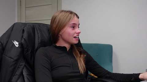 Media: Video of a young, Caucasian woman with long, blonde hair, wearing a black long-sleeved shirt, sitting in a black Nike jacket on a teal chair, indoors.