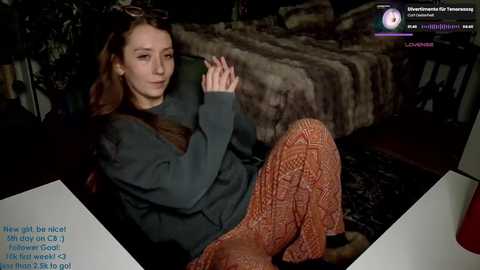 Media: Video of a woman with long brown hair, wearing a dark gray sweater and orange patterned pants, lounging on a plush, dark rug, in a dimly lit room.