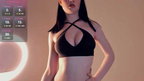 Media: Video of a slim, fair-skinned Asian woman with long black hair, wearing a black halterneck bra that accentuates her medium-sized breasts. She stands against a beige background.