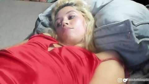 Media: A video of a blonde woman in a red tank top, lying on a bed with gray pillows and beige sheets, looking relaxed.