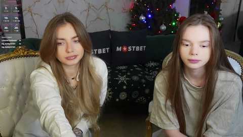 Media: Video of two young women with long, straight brown hair, wearing casual light-colored tops, sitting on a cream-colored sofa. Background features a Christmas tree adorned with colorful lights and decorations.