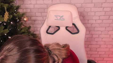 Media: A video of a white gaming chair with a pink brick wall and a decorated Christmas tree in the background, featuring a person with brown hair.