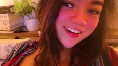 Media: Video of a young woman with long brown hair, light skin, and a warm smile, wearing a colorful striped shirt. Background features a potted plant, books, and a cozy, dimly lit room.