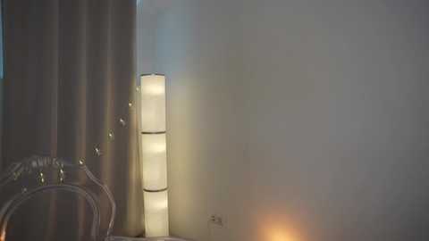 Media: Video of a minimalist room with a cylindrical lamp emitting a soft light, casting a warm glow on a plain white wall. The lamp is on the left, and there's a hint of a metallic object in the background.