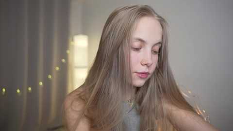 Media: Video of a young Caucasian woman with long, straight, light brown hair, wearing a grey top, looking down thoughtfully. Background features soft, diffused lighting and a blurred, neutral-toned wall.