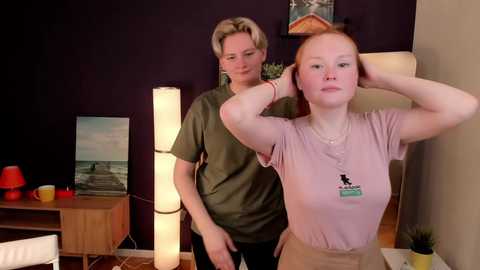 Media: Video of a blonde woman with short hair in a green t-shirt, and a redhead in a pink t-shirt with a badge, both standing in a modern room with a tall floor lamp and artwork.