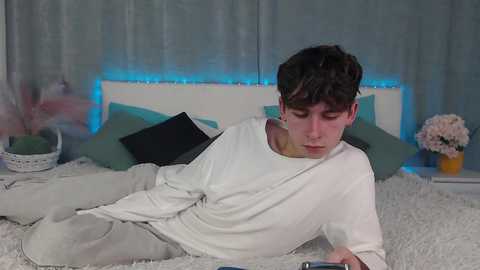 Media: Video of a young man with short brown hair, wearing a white long-sleeved shirt and gray pants, lying on a white shag rug in a dimly lit bedroom with blue LED strip lights, green pillows, and a floral bouquet.