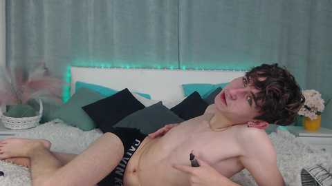 Media: Video of a young, shirtless, fair-skinned man with curly brown hair lying on a bed with white and turquoise pillows. He wears black Calvin Klein underwear, with a relaxed, slightly seductive expression.